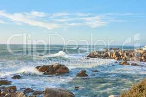 Copyspace at sea with a cloudy blue sky background and rocky coast in Western Cape South Africa. Ocean waves crashing onto boulders at a beach. Peaceful scenic landscape for a relaxing summer holiday