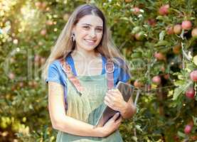 Portrait of a female farmer holding a digital tablet while standing by apple trees. Smiling woman using technology to prepare for harvest on her farm. Monitoring plant growth and agriculture