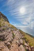 Secluded mountain trail above a beautiful ocean view and cloudy blue sky. Mountainous walking path surrounded by green grass and rocks. Remote destination to explore and enjoy adventure walks