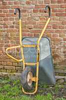 Garden wheelbarrow leaning against a red brick wall in a home backyard. Landscaping equipment and tools to carry and transport soil, manure, compost and fertilizer. Done cleaning a landscaped yard