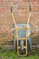 Garden wheelbarrow leaning against a red brick wall in a home backyard. Landscaping equipment and tools to carry and transport soil, manure, compost and fertilizer. Used to lift construction supplies