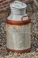 An old rusted metal milk can on the ground from above. An antique dairy canister outside on a pebble covered walkway. A rusty and used metallic jug during the day with copy space