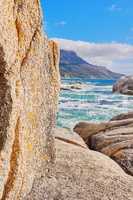 Stunning seaside location for a summer holiday in Cape Town. Boulders at a beach with ocean waves and water washing over rocks at the coast with mountain and blue sky with clouds in the background