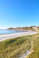 Copyspace at the beach with blue sky background above the horizon. Calm ocean waves across an empty sea along the shore with a grassy meadow. A tranquil landscape for a relaxing summer holiday