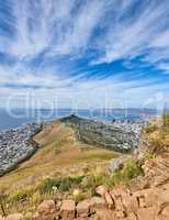 Rocky mountain with view of a costal city by the sea in Cape Town. Landscape of green hill top with stones and plants surrounded by an urban city and the ocean on a cloudy blue sky with copy space.