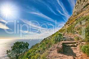 A mountain trail with blue sky and lensflare over ocean. Landscape of mysterious dirt road for hiking on adventure walks along a beautiful scenic trail with lush shrubs in Cape Town, South Africa