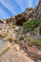 A rocky mountain with plants and shrubs growing against a cloudy sky background with copy space. Rugged, remote and quiet landscape with rocks and stones on a cliff leading to a mysterious cave