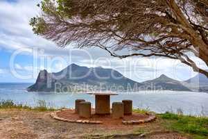 Public bench overlooking the ocean and mountain looks beautiful out in nature. Landscape of four empty chairs and a table with a tree giving them shade offers a relaxing and romantic outdoor sitting