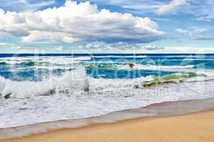 Waves rolling and crashing the shore at the beach during a summer day outside. Sea, ocean current and tide washing onto the seashore under a cloudy blue sky outdoors in nature