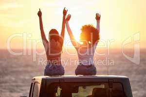 Sing your song to the sun. Rearview shot of two young unrecognizable females celebrating while watching the sunset at the beach.