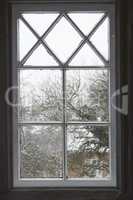 Closeup of a glass window from inside, looking out on cold weather in the morning. A wooden windowpane with bad insulation, shut and closed with a view of a winter scene with snow, frost and ice.