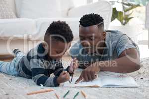 Working on those motor skills. a young father lying on the the living room floor and helping his son draw.