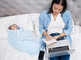 Some peace and quiet. a mother using her laptop while her baby sleeps on the bed.