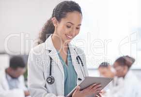 Accessing patient records has never been easier. an attractive young female doctor using a tablet while standing in the boardroom with her colleagues in the background.