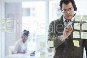 Planning to reach his targets. a young businessman wearing a headset while brainstorming with notes on a glass wall in an office.