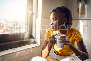 A thinking and coffee drinking black woman enjoys her morning routine while gazing outside a window on a bright sunny day. Beautiful female looks thoughtfully towards city while sitting at home