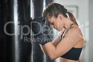 Closeup of a tired and exhausted strong female athlete boxer, after a training session leaning on punching bag in the gym, side view. Fit and active woman taking a rest break after her exercise
