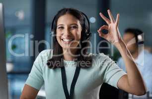 You talk, well take care of it. Portrait of a young woman using a headset and showing an okay gesture in a modern office.