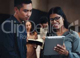 Getting his input. two young businesspeople looking over a tablet in the office with their colleagues in the background.