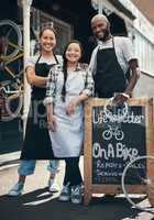 Come, share the journey with us. Portrait of three young workers standing behind a bicycle repair sign outside.