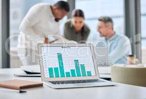 Future proof your business through trend analysis. a laptop with graphs on display in an office with businesspeople working in the background.