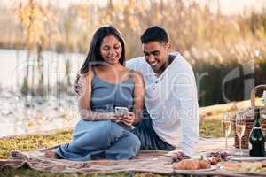 Sharing in a cosy connection. a young couple using a cellphone while on a picnic at a lakeside.