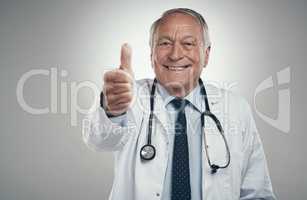 Years later i still love my job. an elderly male doctor in a studio giving a thumbs up against a grey background.