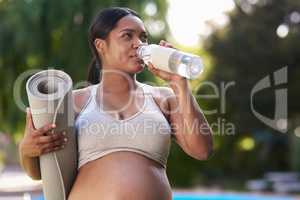 Hydration is essential, especially when youre pregnant. a pregnant woman holding her yoga mat and drinking water while standing outside.