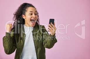 I just won the prize. an attractive young woman standing against a pink studio background and feeling excited while using her cellphone.