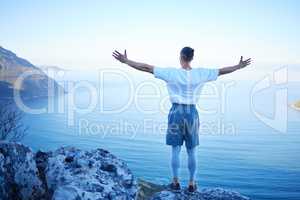 What freedom looks like. Rearview shot of an unrecognizable young man standing with his arms outstretched while looking at the ocean view from the mountain.