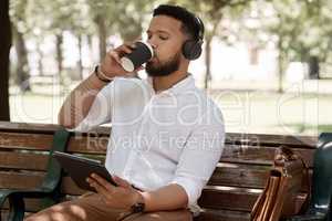 Enjoying a quick coffee break out in the fresh air. a young businessman wearing headphones and using a digital tablet while sitting on a bench outdoors.