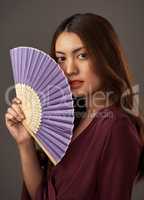 Fan the flames of beauty. Cropped portrait of an attractive young woman posing with a fan in studio against a grey background.