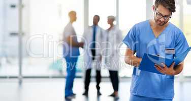 Young male doctor reading, holding and looking at patient health information at a hospital. Medical professional standing with doctors in the background. Healthcare worker working on a clinic file