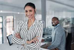 Were on the path to success. an attractive young businesswoman standing with her arms folded while a colleague works behind her.