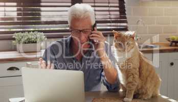 My clients know Im always ready to help them. a senior man sitting alone in the kitchen with his cat and using technology to work from home.