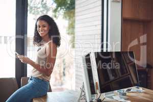 Smiling, modern and casual female in her home office happy to be working remote. Portrait of a confident young online worker sitting on a desk. Digital marketing employee with a smile holding a phone