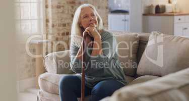 I have all the time in the world to think. a senior woman sitting alone on the sofa at home and looking contemplative while holding her walking stick.