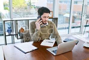 Ensuring all his plans are in order. a young businessman talking on a cellphone while working on a laptop in an office.