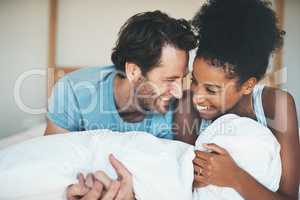 Happy, loving and funny interracial couple smiling and laughing while lying in bed and sharing a tender moment. Carefree husband and wife relaxing and having fun while bonding in the bedroom