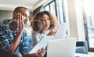 Black couple talking, reading finance papers and planning their bills while working on tablet and laptop on the couch together at home. Husband and wife going through paperwork for tax and loans