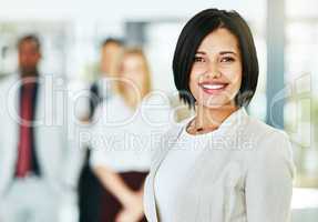 Young, smart and smiling businesswoman with a diverse team at work ready to achieve success. Confident female professional executive standing in a modern office with her colleagues in the background