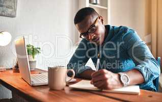 African american entrepreneur writing in a notebook and planning a project strategy. Young man remote working from home with a laptop at his startup business while scheduling appointments in a book