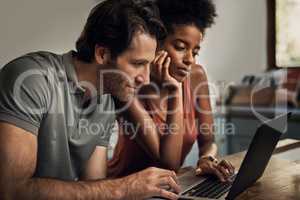 Diverse couple searching for a new rental apartment home on their laptop sitting together in their kitchen at their house. Man and woman in an interracial marriage relationship doing online shopping