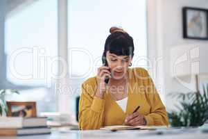 This is 21st century business. attractive young businesswoman sitting alone in her office talking on her cellphone while writing notes.