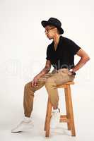 Is this pose fine. Studio shot of a handsome young man wearing a hat and glasses while being seated on a chair against a grey background inside of a studio.