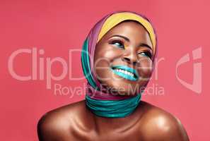 Feeling vividly blissful. Studio shot of a beautiful young woman smiling while wearing a head wrap and make up against a pink background.