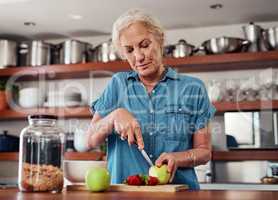 These days, she only eats healthy. an attractive senior woman chopping up apples and other fruit while preparing breakfast in the kitchen.