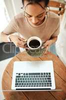 Coffee gets me through my workload. High angle shot of an attractive young businesswoman sitting and enjoying a cup of coffee while using her laptop.