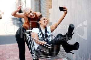 Hashtag fun in the trolley. two cheerful young girlfriends taking a selfie while playing around with a shopping cart outdoors.