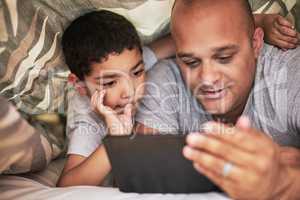 The show has their full attention. a cheerful young man and his son watching videos on a digital tablet together while hanging out on a bed at home during the day.